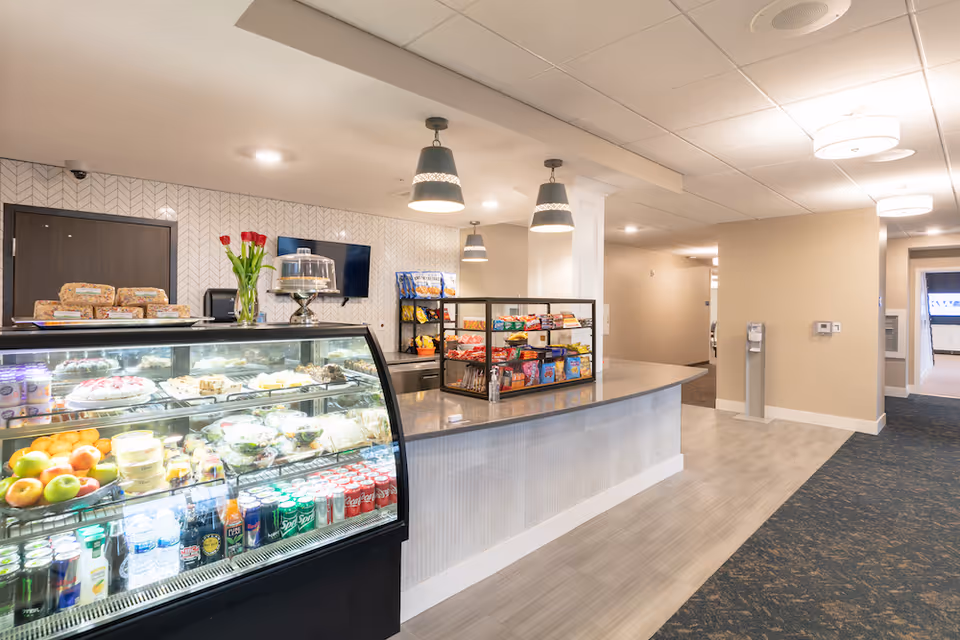 Interior view of a snack and beverage counter area in a senior living facility. The counter has a glass display case filled with fruits, packaged salads, desserts, and various canned and bottled drinks. Behind the counter, shelves hold bags of chips and other snacks. The area is well-lit with hanging pendant lights and has a modern, clean design with light-colored walls and flooring.
