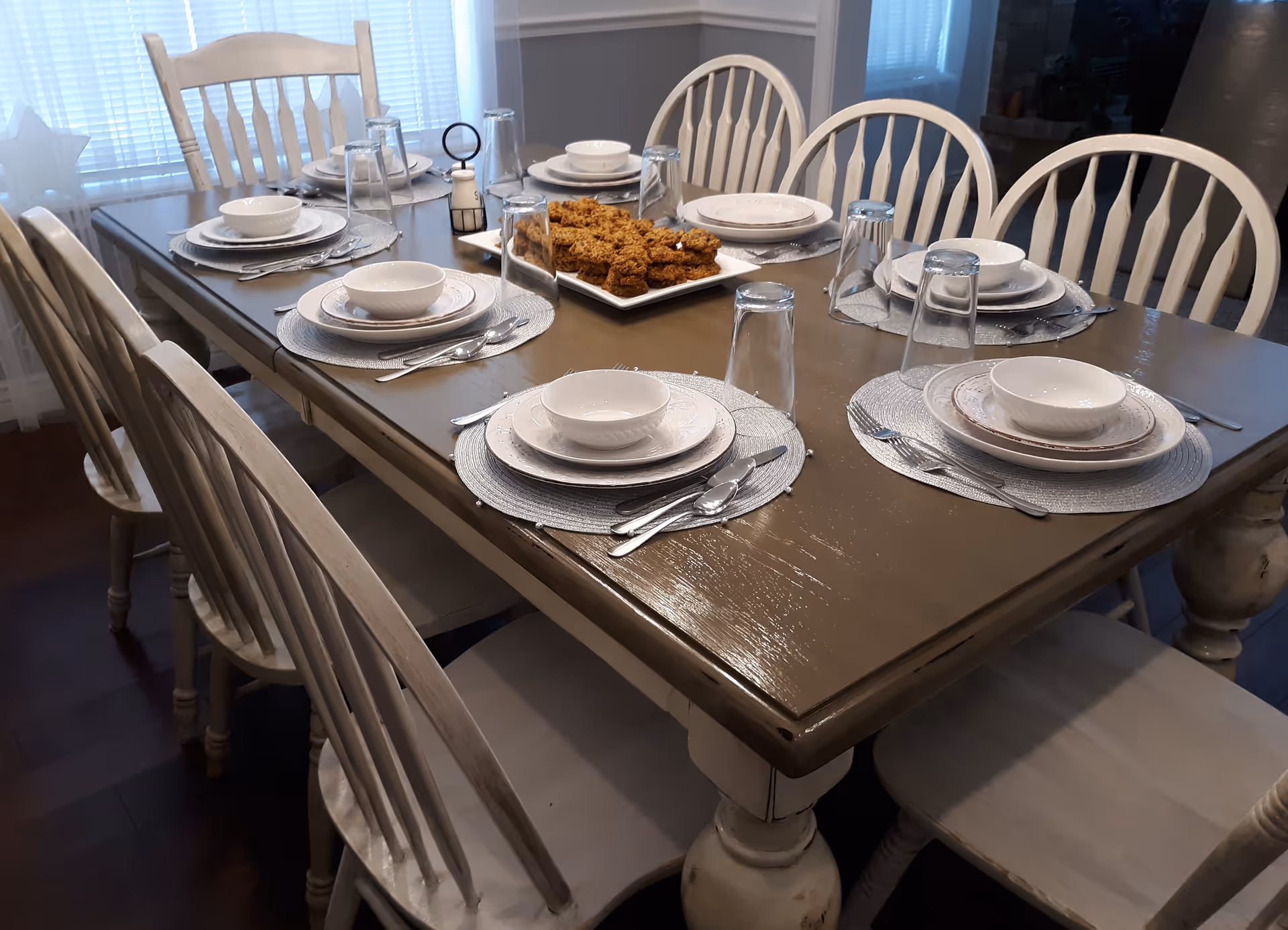 A dining table set for six with white plates, bowls, silverware, and upside-down drinking glasses on silver placemats. A rectangular plate with baked goods is placed in the center of the table. The table is surrounded by white wooden chairs and is positioned near windows with blinds.