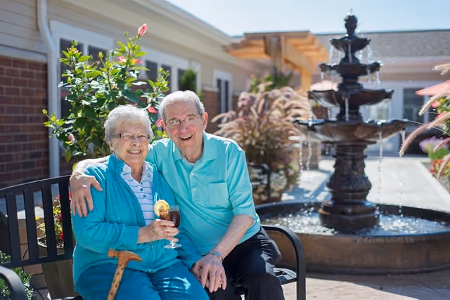 An elderly couple sitting on a bench outdoors near a water fountain. The woman is holding a drink with a lemon slice and a cane rests beside her. Both are smiling and appear happy, with greenery and a building in the background.