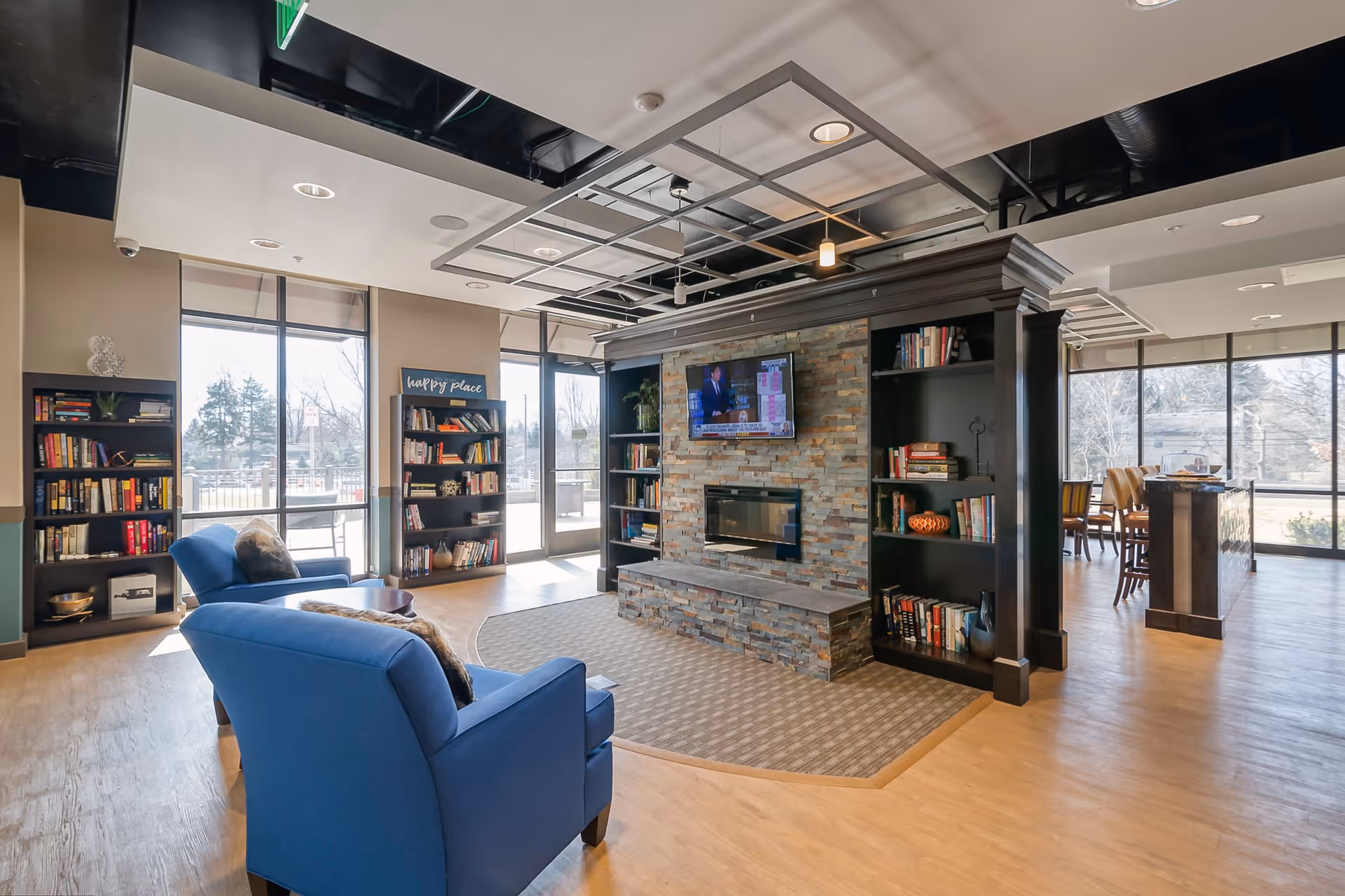 A cozy living room area in a senior living facility featuring two blue armchairs facing a stone fireplace with a mounted flat-screen TV above it. The fireplace is flanked by built-in black bookshelves filled with books and decorative items. Large windows and glass doors let in natural light and offer views of the outdoor area. A dining area with tables and chairs is visible in the background.