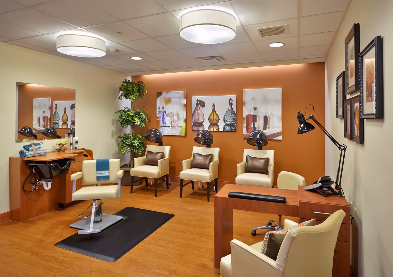 A well-lit salon room with a styling chair in front of a mirror and sink, three cream-colored chairs with brown pillows under hair dryers against an orange accent wall, a wooden desk with a black lamp and phone, and framed artwork on the walls.