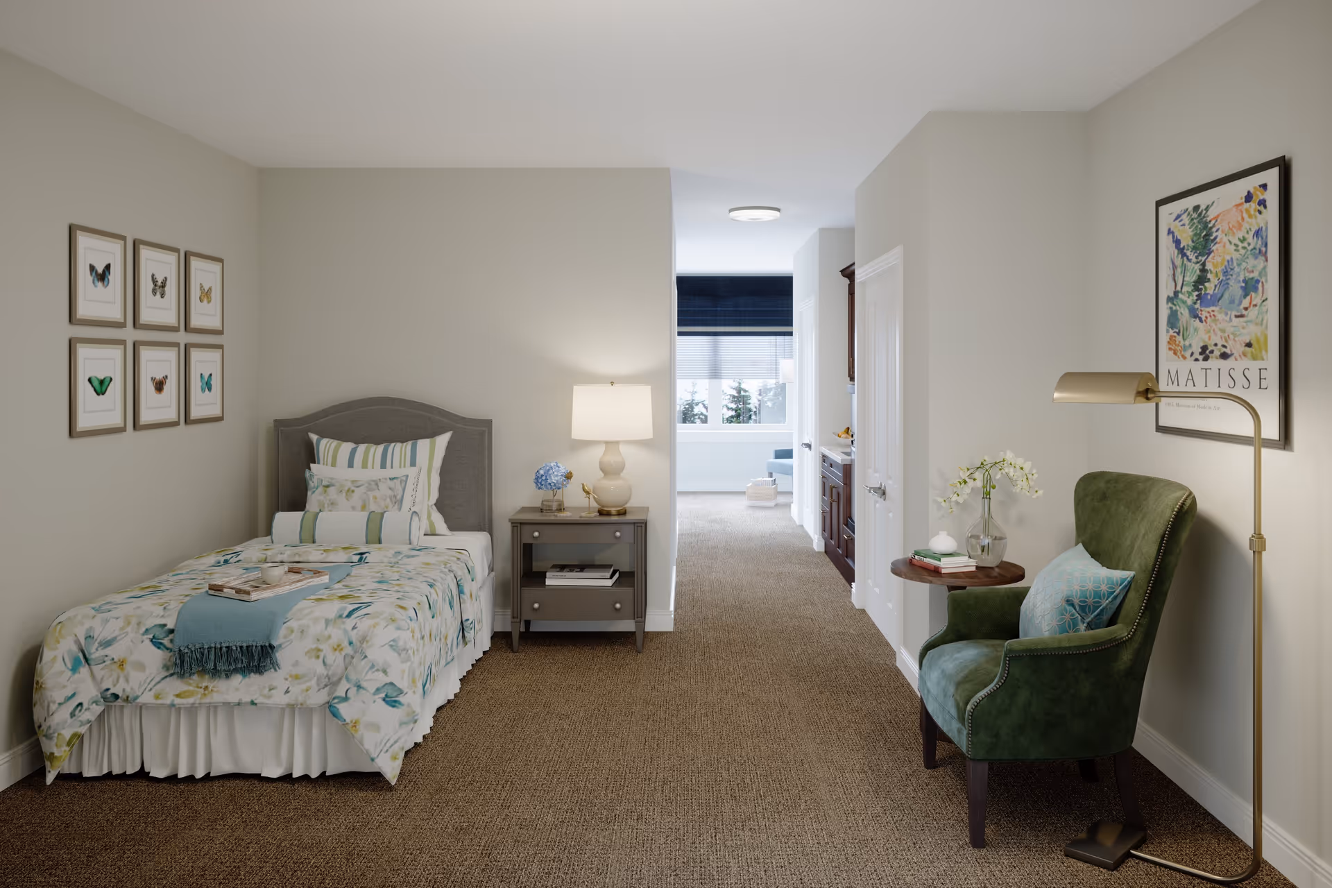 A cozy bedroom in a senior living facility with a single bed adorned with floral bedding and multiple pillows. Next to the bed is a nightstand with a lamp and decorative items. On the right side, there is a green upholstered armchair with a blue cushion, a small round wooden side table with a vase of flowers, and a tall floor lamp. The walls are light-colored with framed butterfly artwork on the left and a Matisse art print on the right. A hallway leads to another room with a window and seating area visible in the background.
