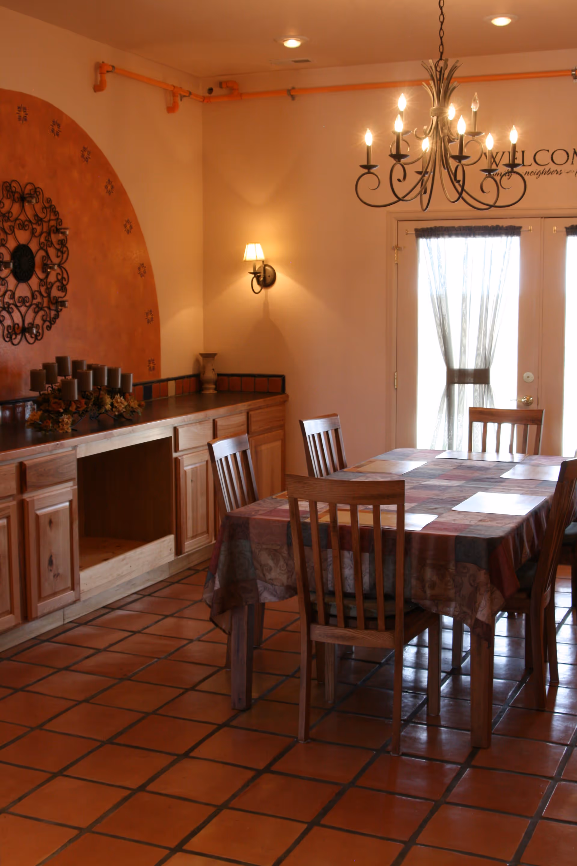 Dining room with a wooden table and chairs, tiled floor, sideboard, chandelier and French doors with sheer curtains.