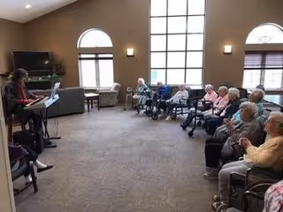 A group of elderly people seated in a semi-circle in a spacious room with large windows, listening to a woman playing an electronic keyboard.