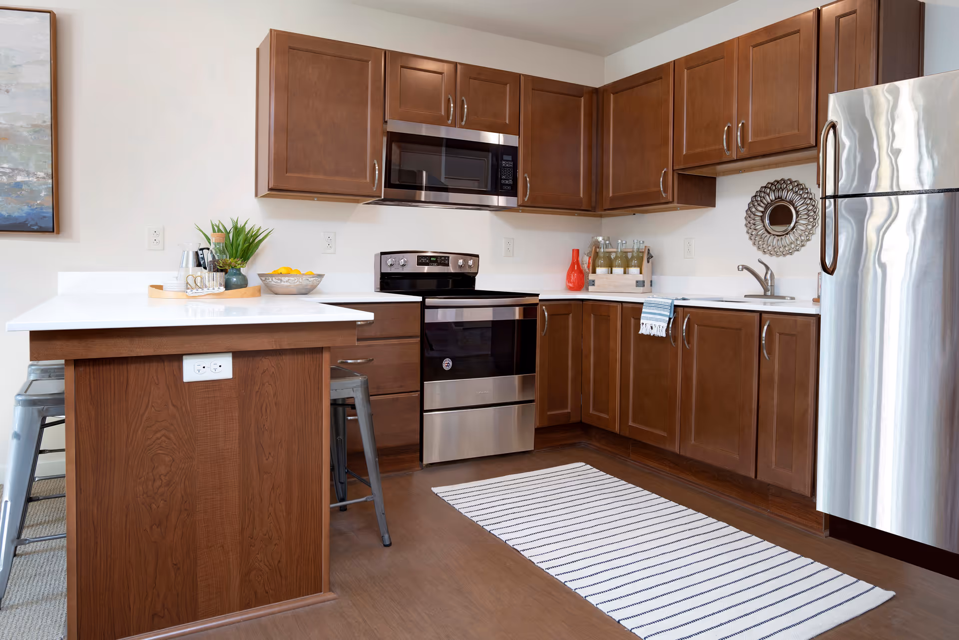 Modern kitchen with wooden cabinets, stainless steel appliances including a refrigerator, oven, and microwave. White countertops with a small island featuring two metal stools. Decorative items include a bowl of lemons, a plant, a red vase, and bottles on the counter. A striped rug is placed on the wooden floor.