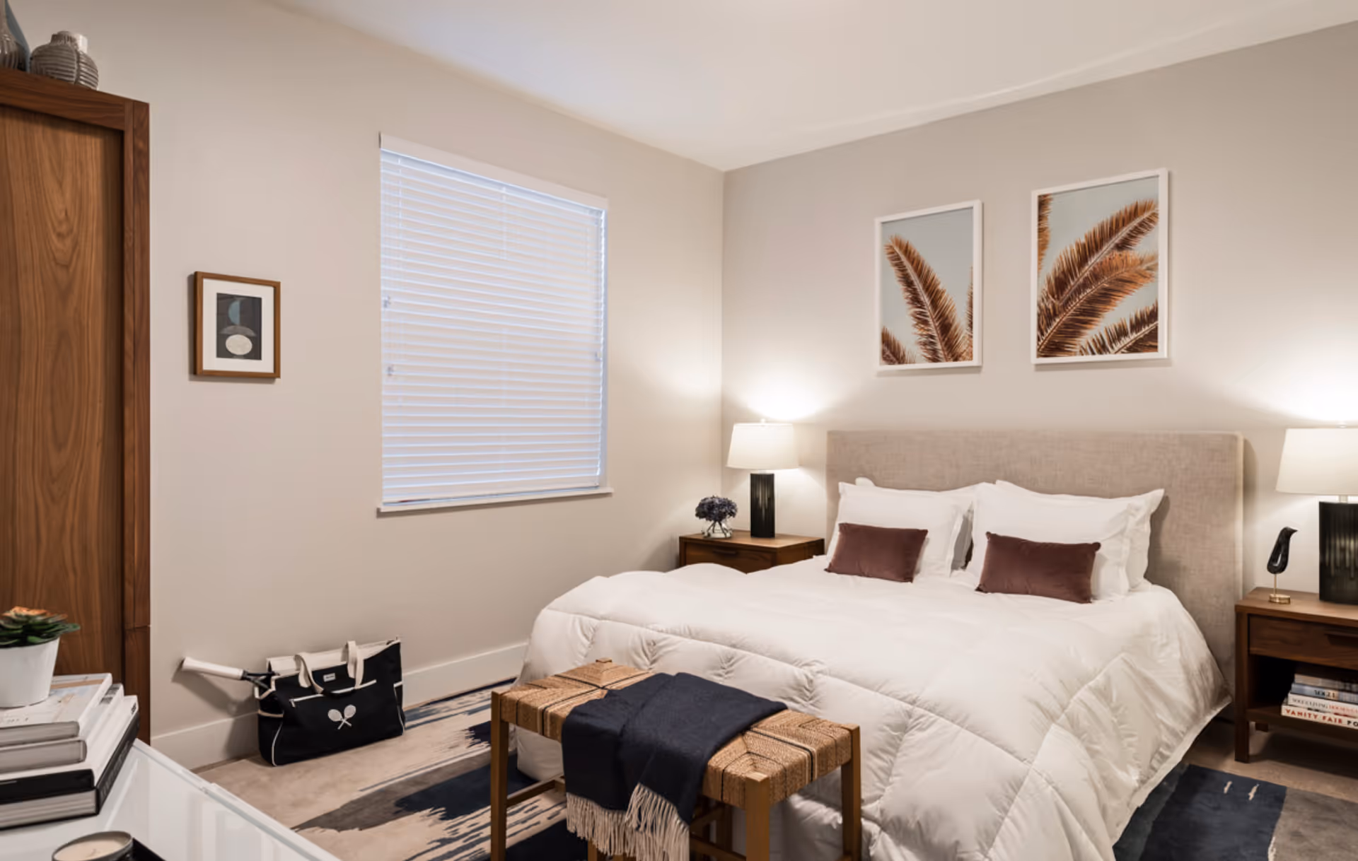 A cozy, modern bedroom with a neatly made bed, nightstands with lamps, framed wall art, and a window with blinds.
