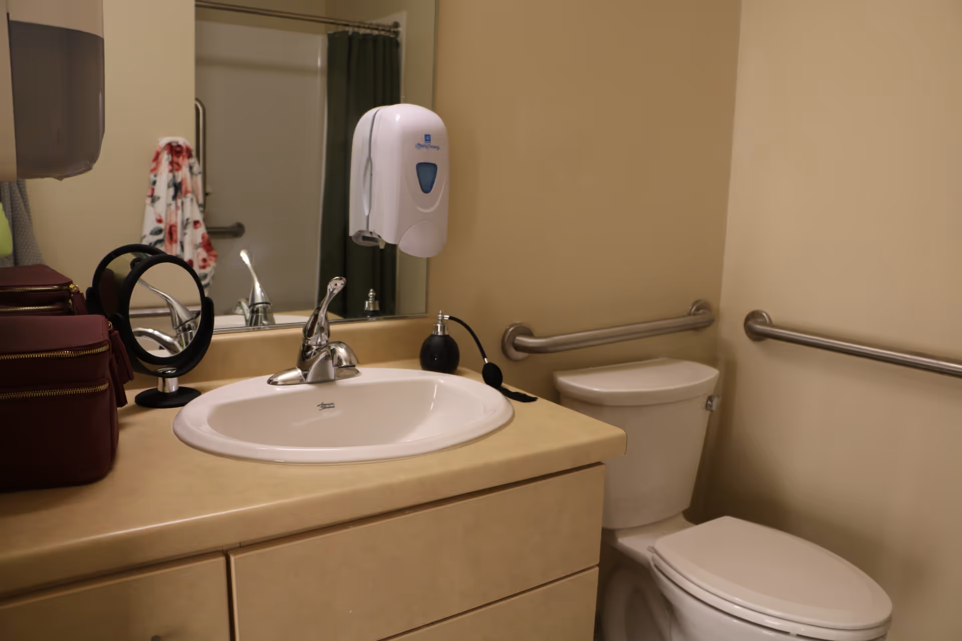 A bathroom with a beige countertop featuring a white sink and silver faucet. There is a toilet with safety grab bars on the walls beside it. A large mirror is mounted above the sink, reflecting a shower area with a green curtain and a floral towel hanging on a grab bar. A soap dispenser is attached to the wall above the sink, and a small round mirror and a maroon toiletry bag are on the countertop.