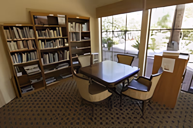 A small library or reading room with a wooden table surrounded by four chairs. There are bookshelves filled with books along the left wall and large windows on the right side letting in natural light and showing some greenery outside.