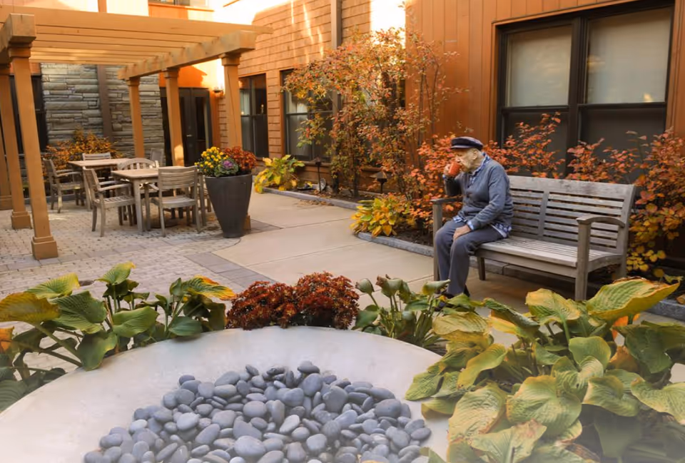 An elderly man wearing a hat and blue sweater sits on a wooden bench in a courtyard, drinking from a red cup. The courtyard features a stone fire pit surrounded by green and yellow plants, a wooden pergola with tables and chairs, and autumn-colored bushes against the building walls.
