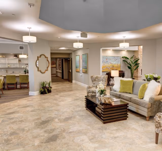 Well-lit senior living facility common area with sofas, armchairs, a coffee table, and a dining area in the background.