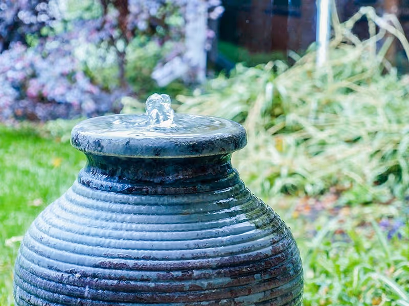 A close-up view of a ceramic water fountain with water gently bubbling from the top, set against a blurred background of green grass and plants in an outdoor garden area.