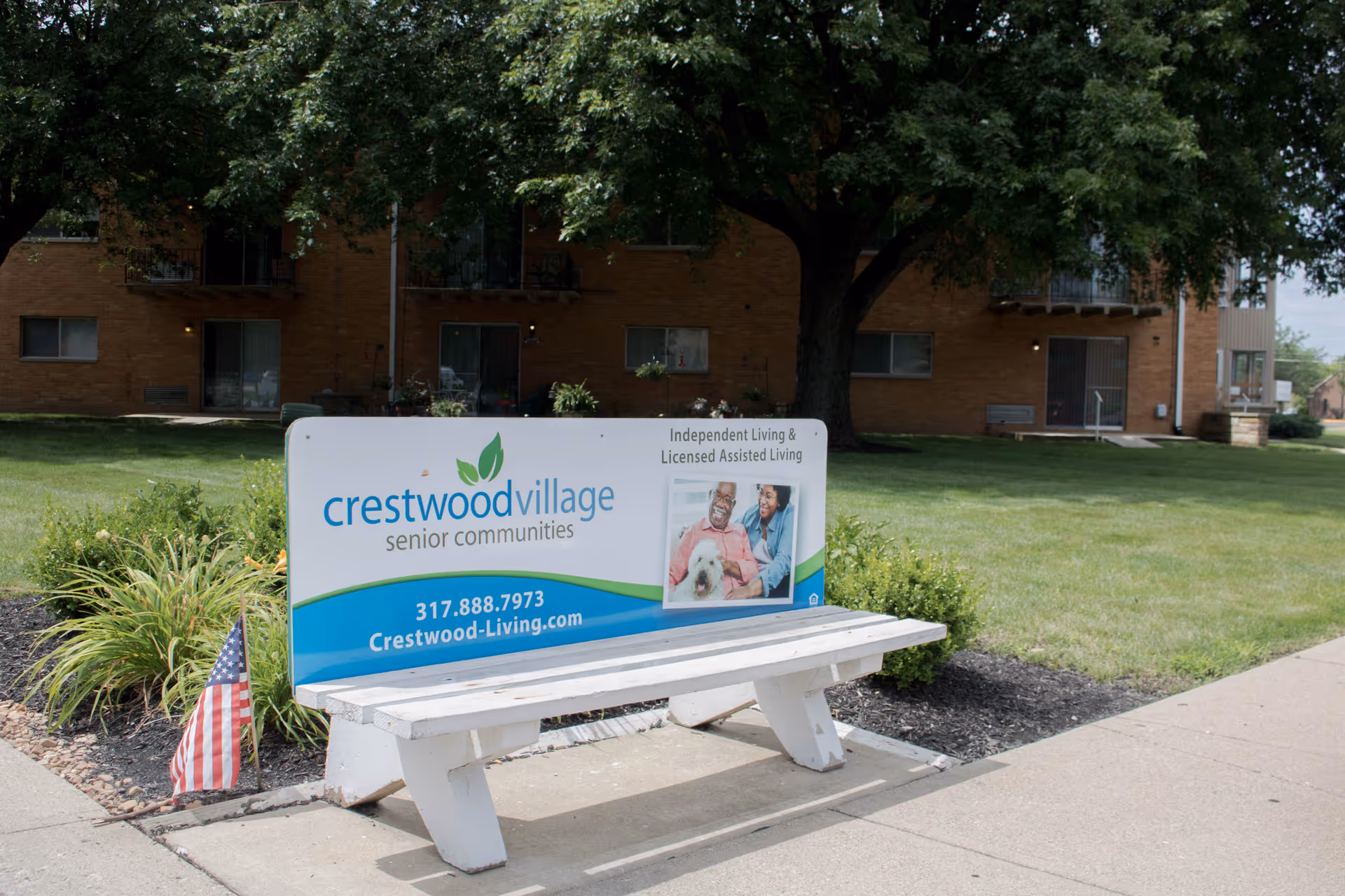 Outdoor view of Crestwood Village Senior Communities sign placed on a white bench in front of a grassy area with trees and a brick building in the background. The sign includes contact information and a photo of two seniors with a dog.