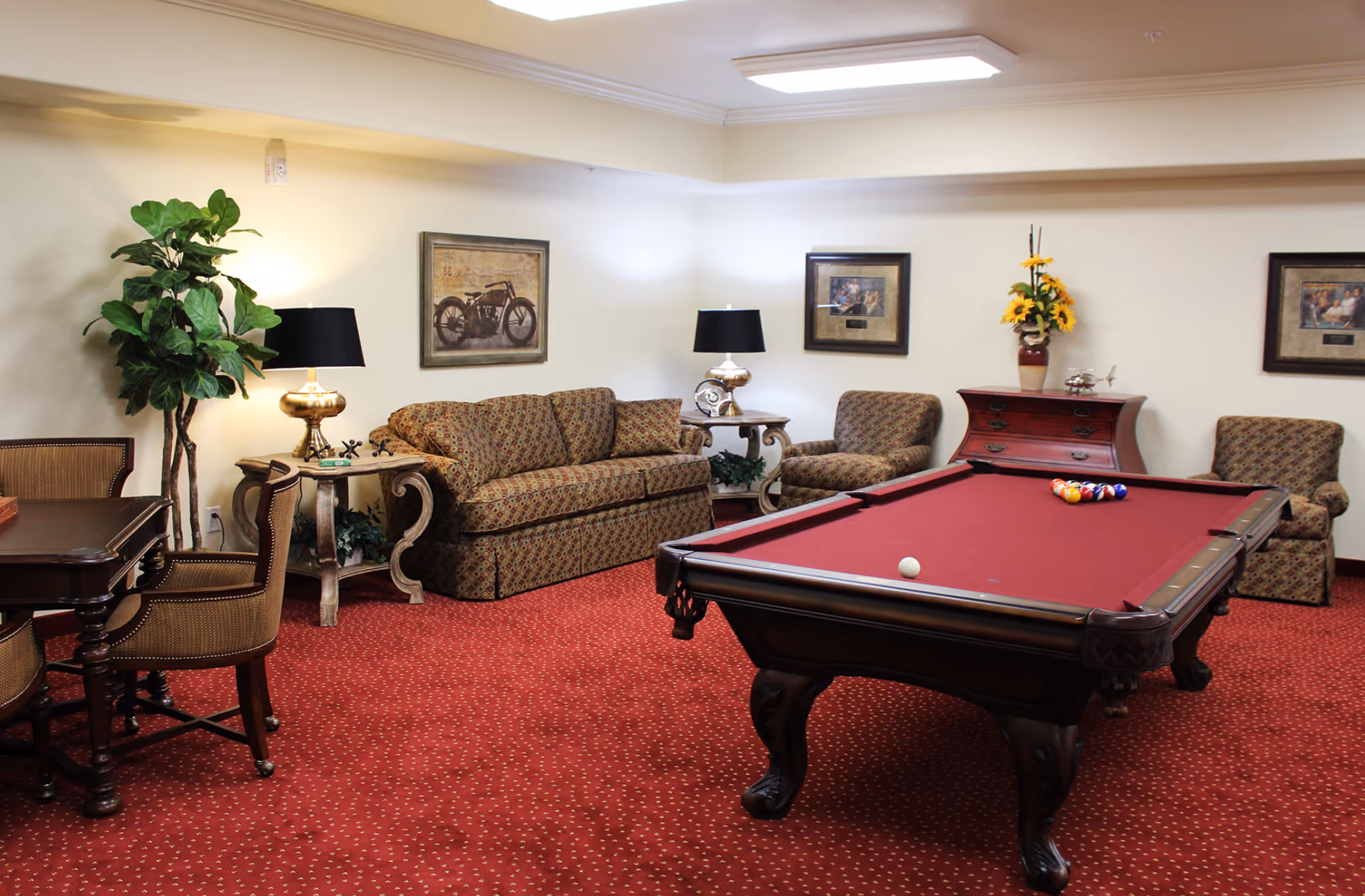 A cozy common room with a red-felt pool table, patterned sofa and chairs, side tables with lamps, and framed artwork on the walls.