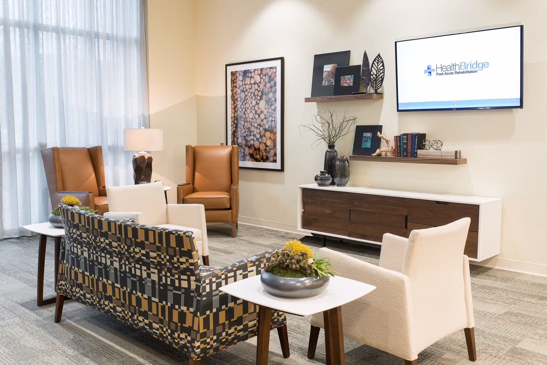 A modern and cozy living room area with a patterned sofa, two white armchairs, and two brown leather armchairs. There are side tables with decorative plants and a lamp. On the wall, there is a large framed picture of stacked wood logs, two floating shelves with decorative items and books, and a flat-screen TV displaying the HealthBridge Post-Acute Rehabilitation logo. The room has large windows with sheer curtains allowing natural light to enter.