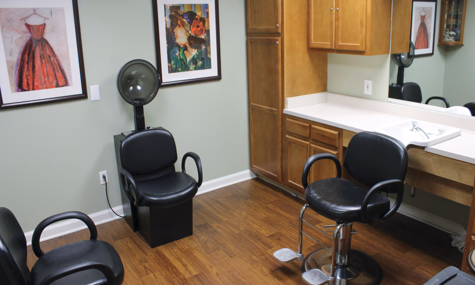 Small hair salon room with styling chairs, a sink and cabinets, and framed artwork on the walls.