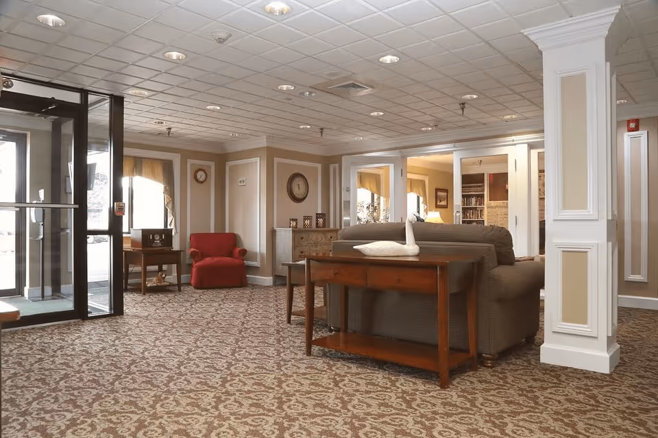 Interior view of a senior living facility lobby with patterned carpet, a glass entrance door on the left, a red armchair near a window, a wooden table with a decorative swan towel on a sofa, and beige walls with white trim and a clock.