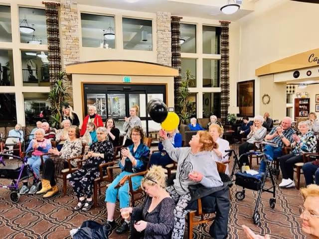 A group of elderly people seated in a spacious, well-lit common area with large windows and stone pillars. Some individuals are clapping, and one woman is holding black and yellow balloons. The room has patterned carpet and a welcoming atmosphere.
