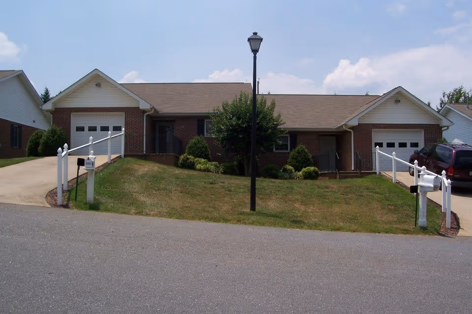 Front exterior view of a single-story brick residential building with two garages, two driveways, a central lawn with a streetlamp, and mailboxes at the edge of the road under a partly cloudy sky.