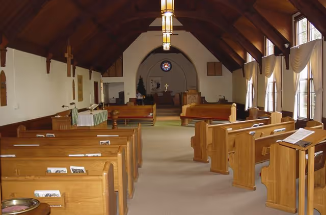 Interior view of a chapel-like room with wooden pews, a vaulted wood ceiling, and an altar at the front.