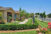 View of a single-story senior apartment building with a well-maintained lawn, shrubs, and flowers along a sidewalk and a street with streetlights in the background under a clear blue sky.