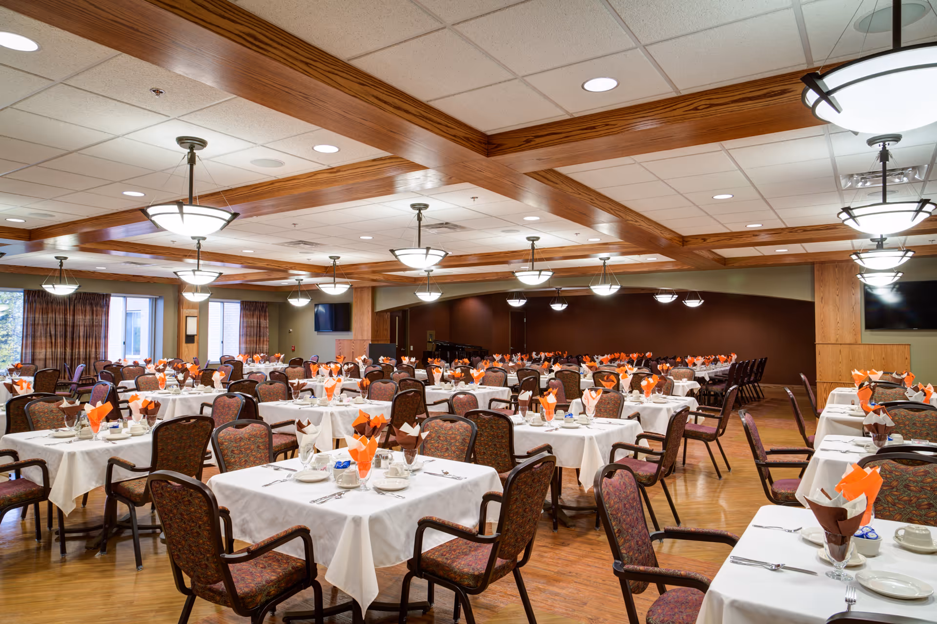 A large dining room with multiple tables covered in white tablecloths, each set with plates, cups, silverware, and orange and brown folded napkins. The room features wooden beams on the ceiling, hanging light fixtures, patterned upholstered chairs, and large windows with curtains allowing natural light to enter.
