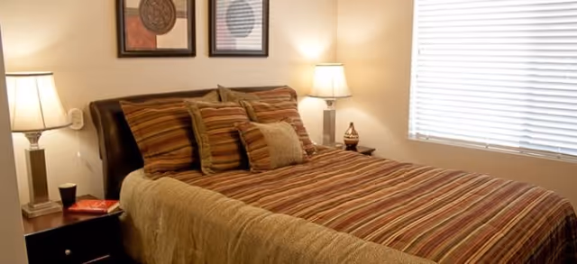 A neatly made bedroom with a striped bedspread, several throw pillows, two bedside tables with lamps, framed wall art, and a window with blinds.