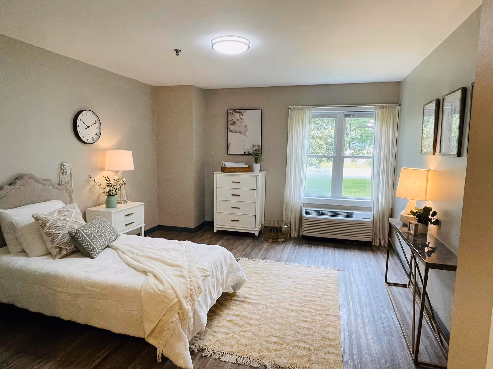 A cozy bedroom in Hanover Manor Assisted Living featuring a single bed with white bedding and decorative pillows, a nightstand with a lamp and a small plant, a white chest of drawers with a basket and plant on top, a large window with sheer curtains, a wall clock, and a console table with a lamp and framed pictures above it. The room has wood flooring and neutral-colored walls.