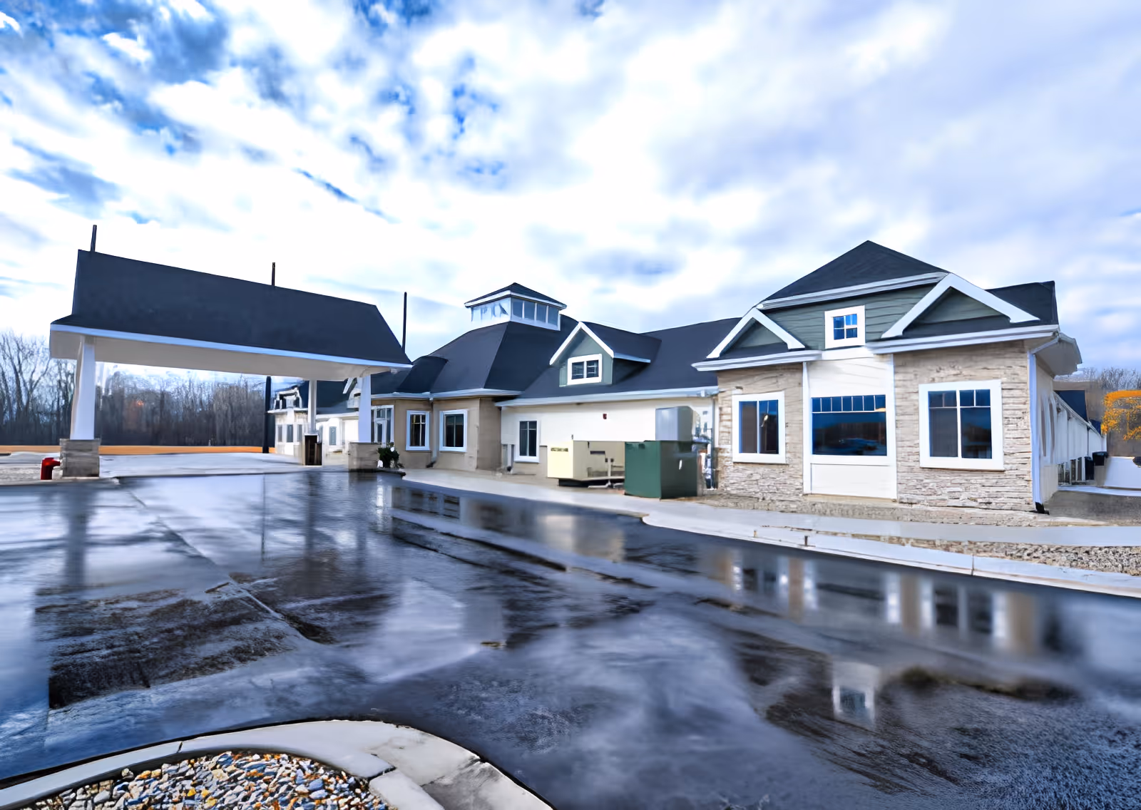 Exterior view of a single-story senior living facility building with a covered entrance driveway, multiple windows, and a mix of stone and siding on the facade under a partly cloudy sky.