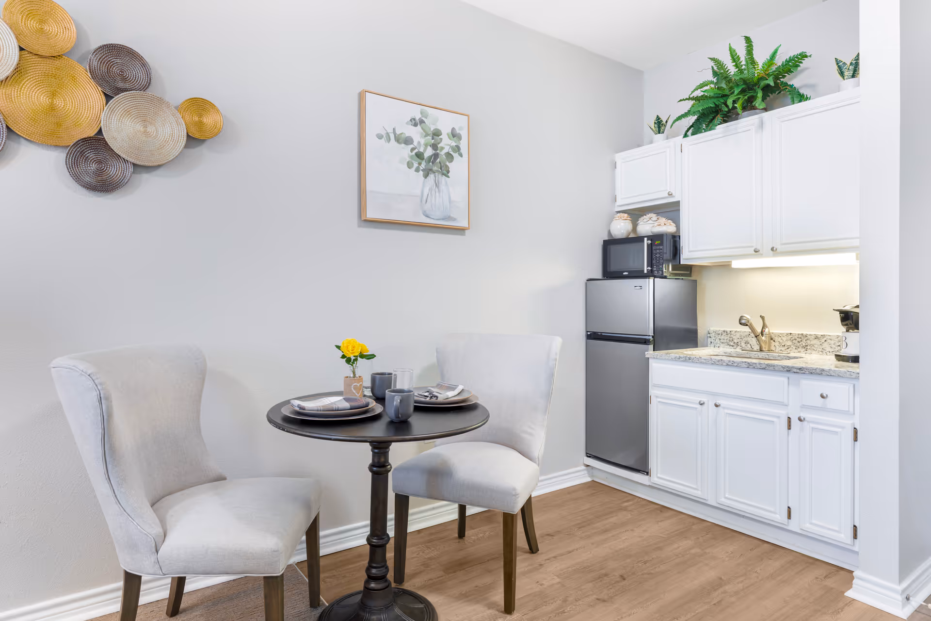 A small dining area with a round black table set for two with plates, mugs, and a small vase with a yellow flower. Two light gray upholstered chairs are placed around the table. The dining area is adjacent to a compact kitchen with white cabinets, a granite countertop, a stainless steel mini refrigerator, and a microwave. Decorative plants and wall art are visible on the walls and above the cabinets.