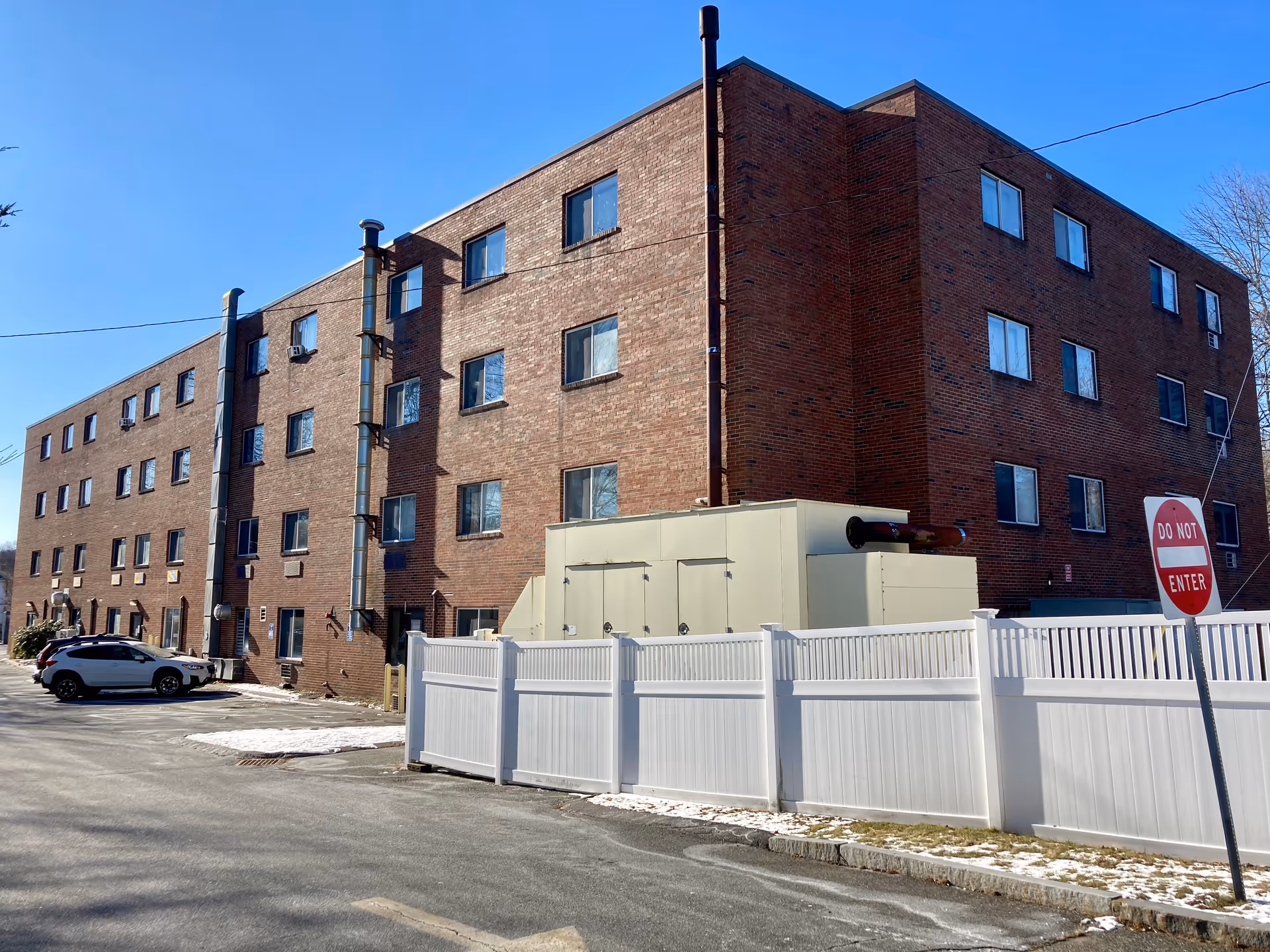 Exterior view of a multi-story brick building with several windows, a white fence in the foreground, a 'Do Not Enter' sign, and a parked car on the left side under a clear blue sky.