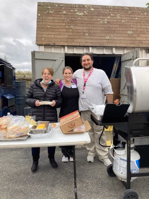 Three people standing outdoors in front of a small shed with open double doors. They are smiling and posing behind a table with food items including buns, cheese slices, and a box of utensils. There is a grill with a propane tank to the right side of the image.