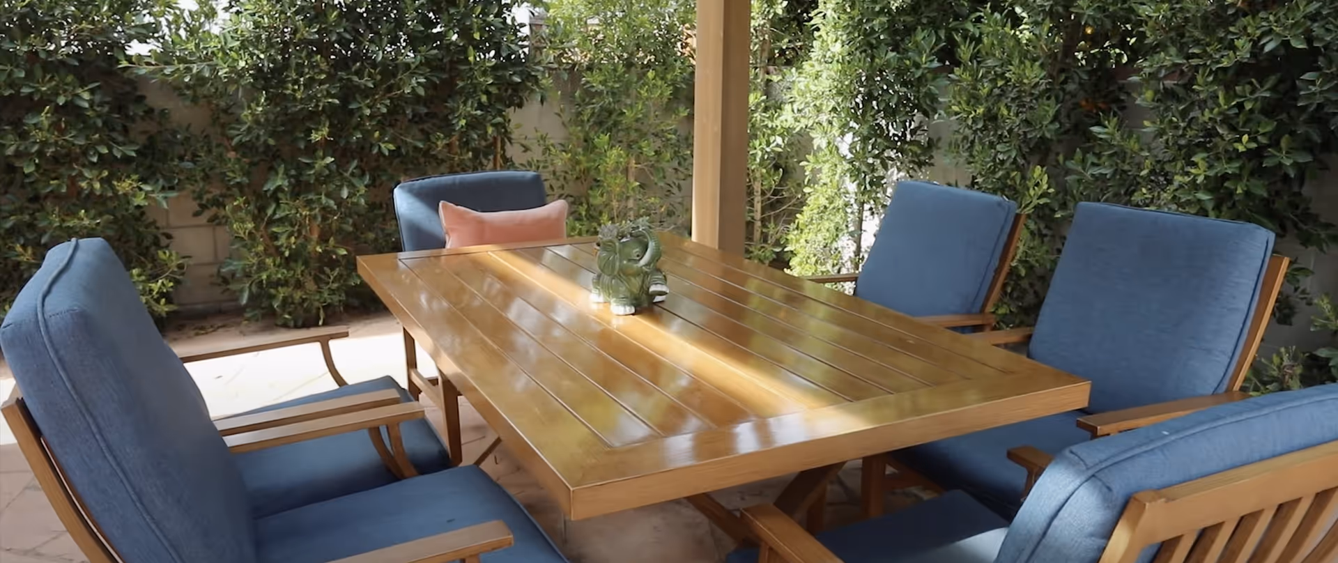 Outdoor patio area with a wooden table surrounded by four blue cushioned chairs. A small green elephant-shaped planter with succulents is placed on the table. The background features dense green foliage and a partial view of a brick wall.