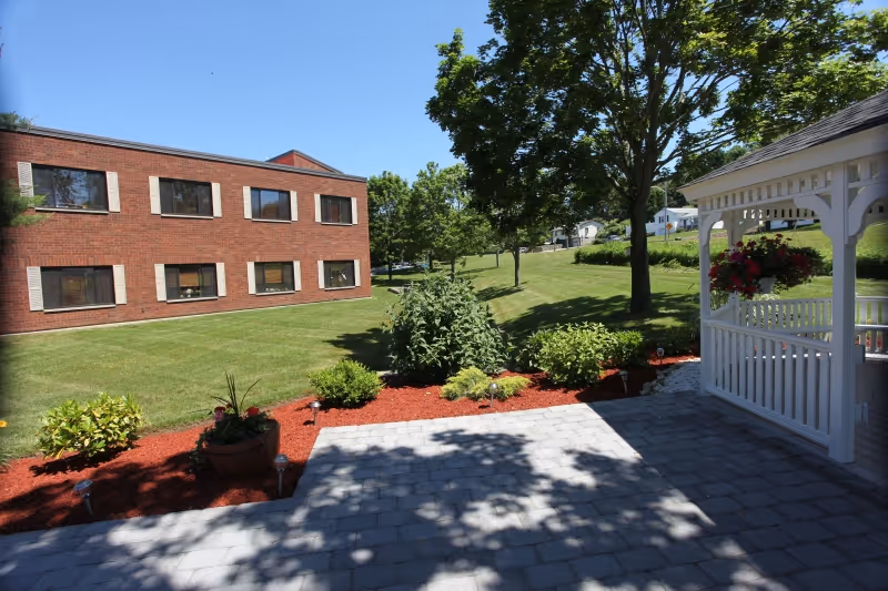Outdoor patio area with a white gazebo on the right, surrounded by landscaped bushes and plants with red mulch. A brick building with multiple windows is visible on the left, and green grass and trees fill the background under a clear blue sky.