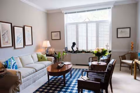 A bright senior living room with a sofa, chairs, flowers, and a large window.