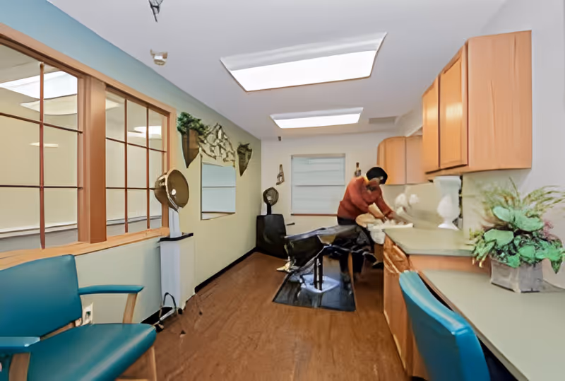 Interior of a hair salon area in a senior living facility with a person seated at a hair washing station. The room has wooden cabinets, a countertop with a plant and decorative items, teal chairs, and large ceiling lights. There is a window with wooden framing on the left wall and a mirror on the far wall.