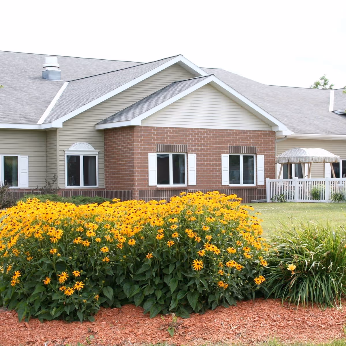 Exterior view of a single-story assisted living facility building with beige siding and red brick accents. The building has multiple windows with white shutters and a gray shingled roof. In front of the building, there is a large flower bed filled with bright yellow flowers and green foliage, bordered by red mulch. To the right, there is a white fence enclosing a small patio area with a canopy.