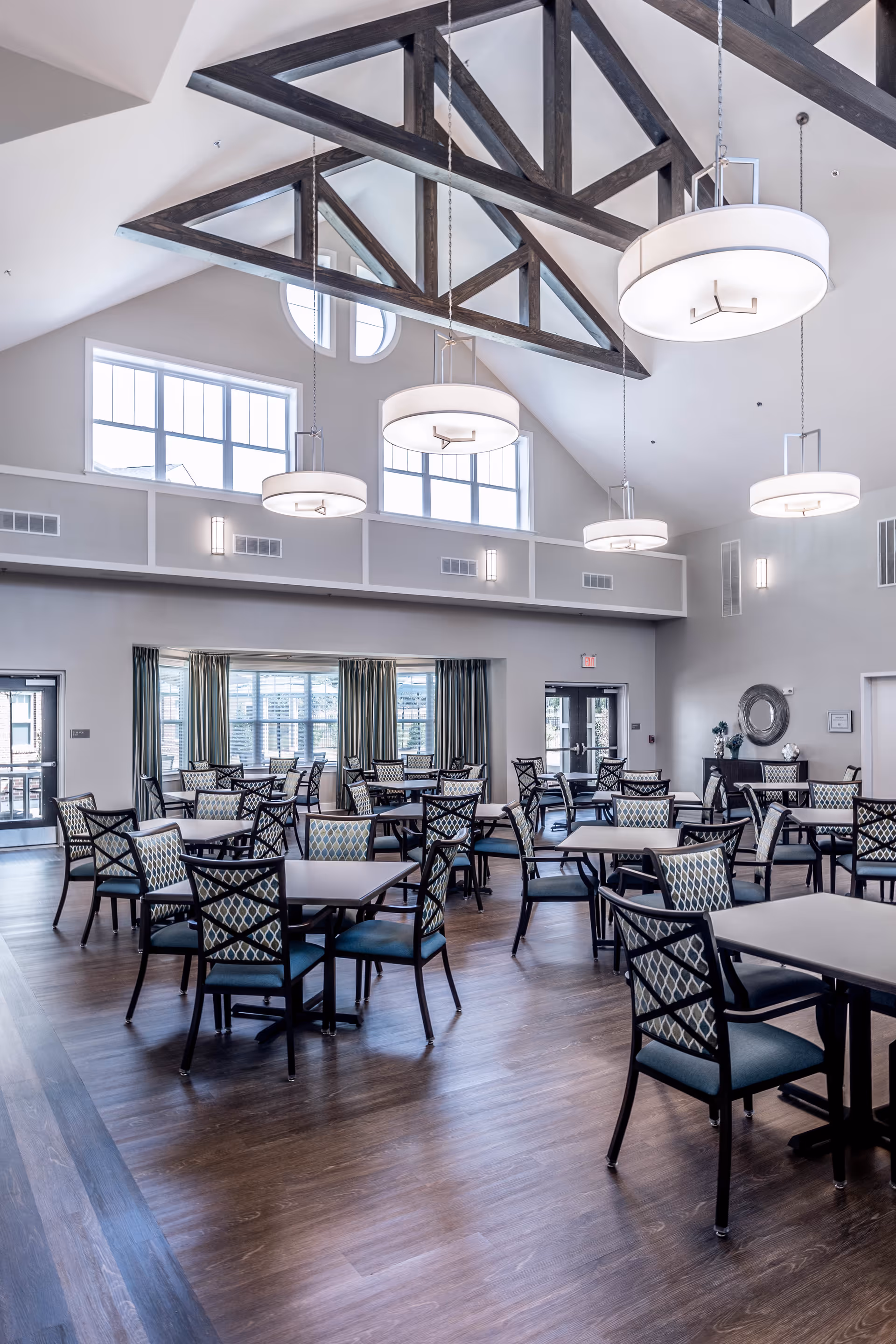 A spacious dining room with multiple square tables and patterned chairs arranged neatly on a wooden floor. The room features high vaulted ceilings with exposed dark wooden beams and large circular hanging light fixtures. There are several windows letting in natural light, and the walls are painted in a light neutral color.