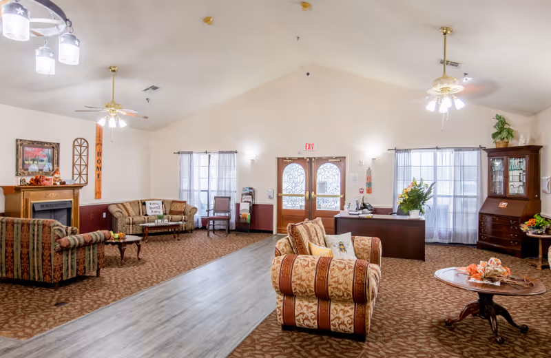 A spacious assisted living facility common area with patterned carpet and a wood-like floor pathway. The room features multiple upholstered armchairs and sofas arranged around small tables. There is a fireplace with decorative items on the mantel, a reception desk with flowers, and large windows with sheer curtains allowing natural light. Ceiling fans and light fixtures hang from the vaulted ceiling.