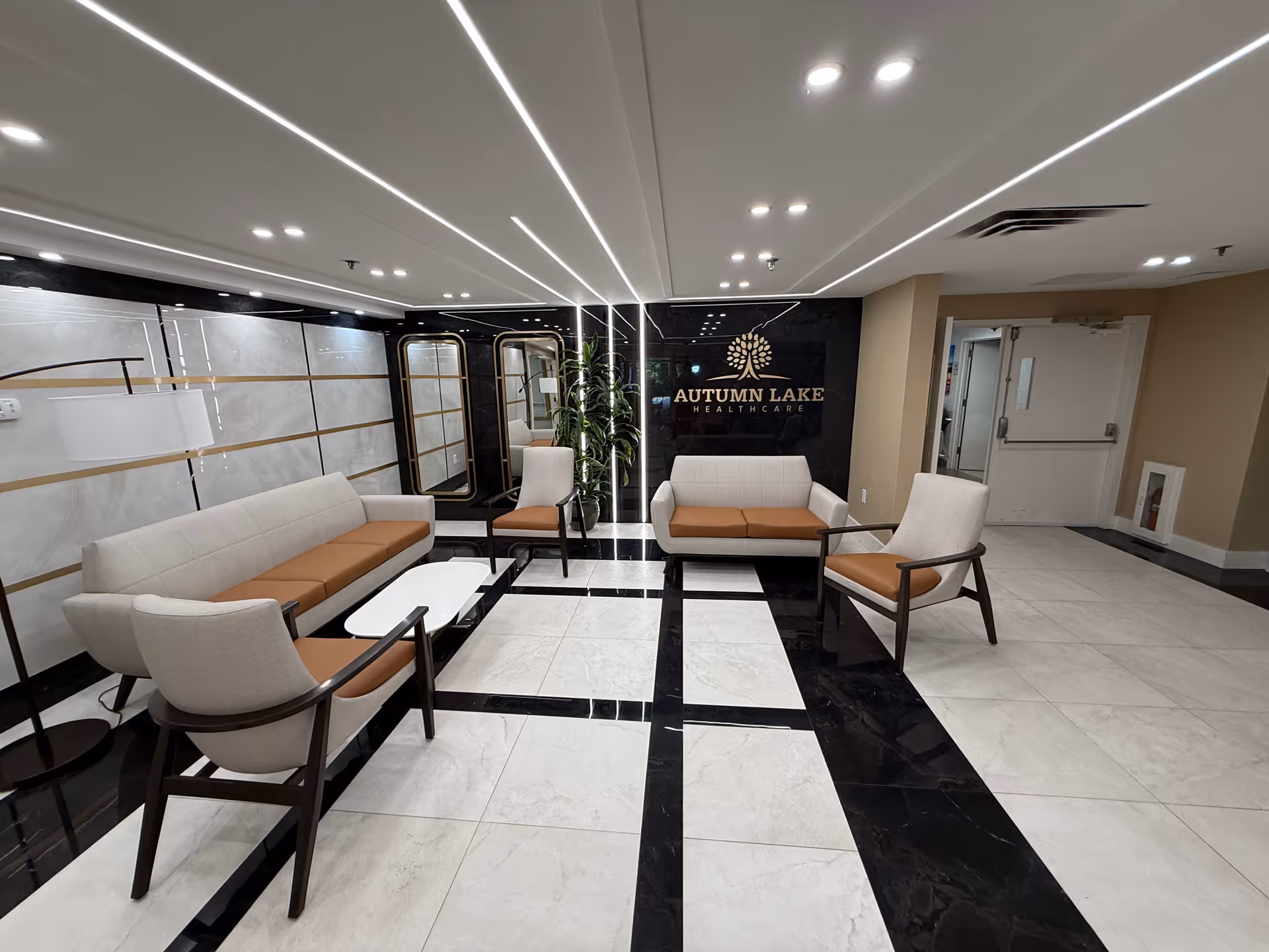 Modern waiting area with beige and brown cushioned chairs and sofas arranged around a white coffee table. The floor has a black and white tile pattern. The back wall features a large black panel with the Autumn Lake Healthcare logo and name. There are mirrors and a tall plant along the left side wall, and a door is visible on the right side.