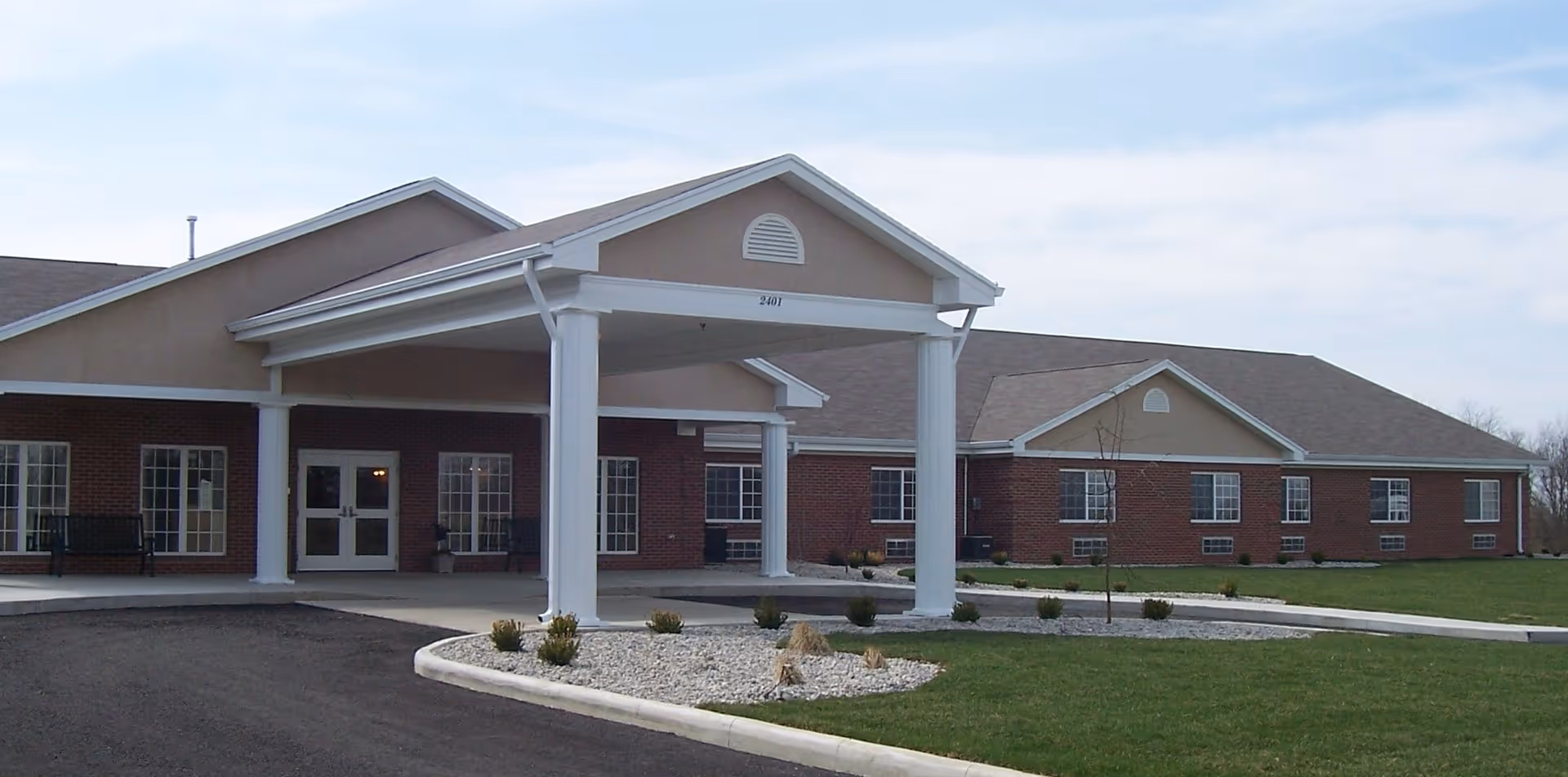 Exterior view of Forest Park Health Campus showing a single-story brick building with a covered entrance supported by white columns, surrounded by a paved driveway, landscaped areas with small shrubs, and a grassy lawn under a partly cloudy sky.