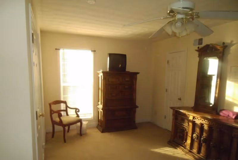 Sparsely furnished bedroom with antique wooden dressers, a mirror, a chair, a small TV on an armoire, and a ceiling fan.