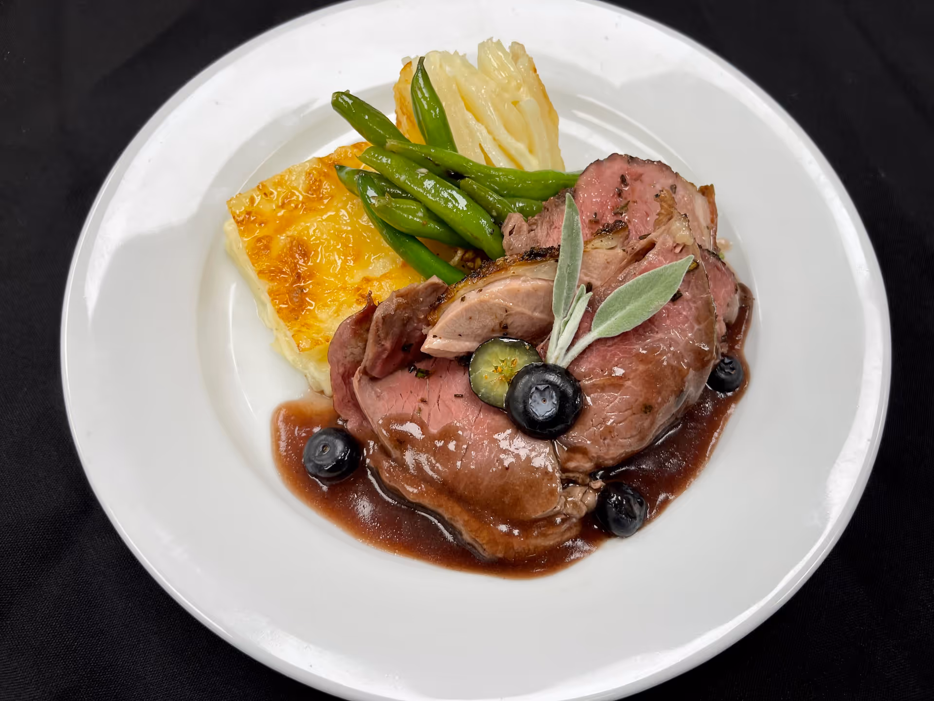 A white plate with slices of roasted meat topped with a dark sauce, garnished with blueberries and a sprig of sage. The plate also contains green beans and a serving of scalloped potatoes, all placed on a black tablecloth.