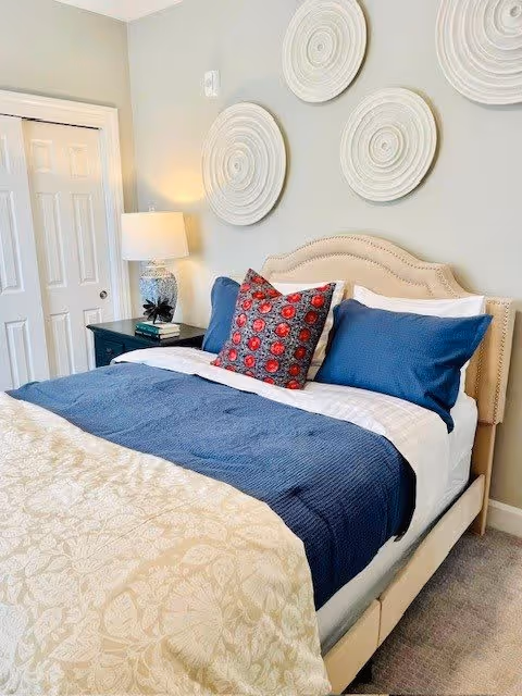 Well-lit bedroom with a beige upholstered bed dressed in blue and white bedding, a patterned accent pillow, a nightstand with a lamp, and circular wall art above the headboard.