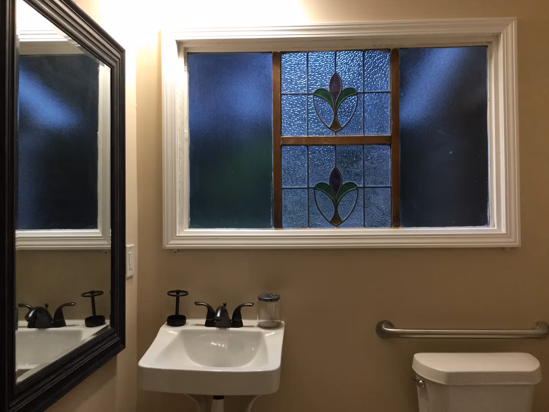 Small bathroom with a pedestal sink, mirror, toilet, grab bar, and a decorative stained-glass window.