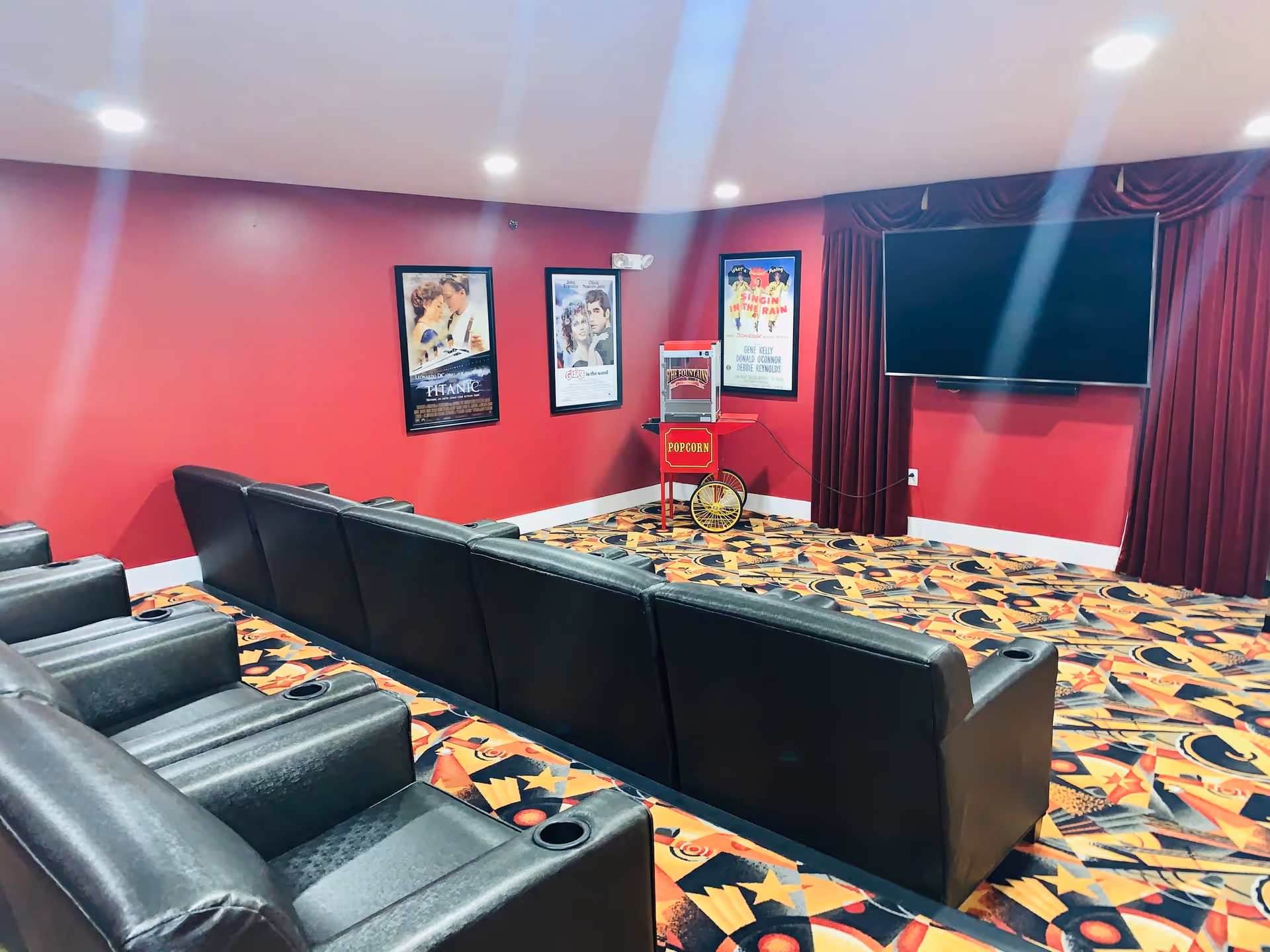 A small movie theater room with red walls and patterned carpet. There are two rows of black leather recliner seats facing a large flat-screen TV mounted on the wall. A popcorn machine is placed in the corner, and three framed classic movie posters are hung on the walls.