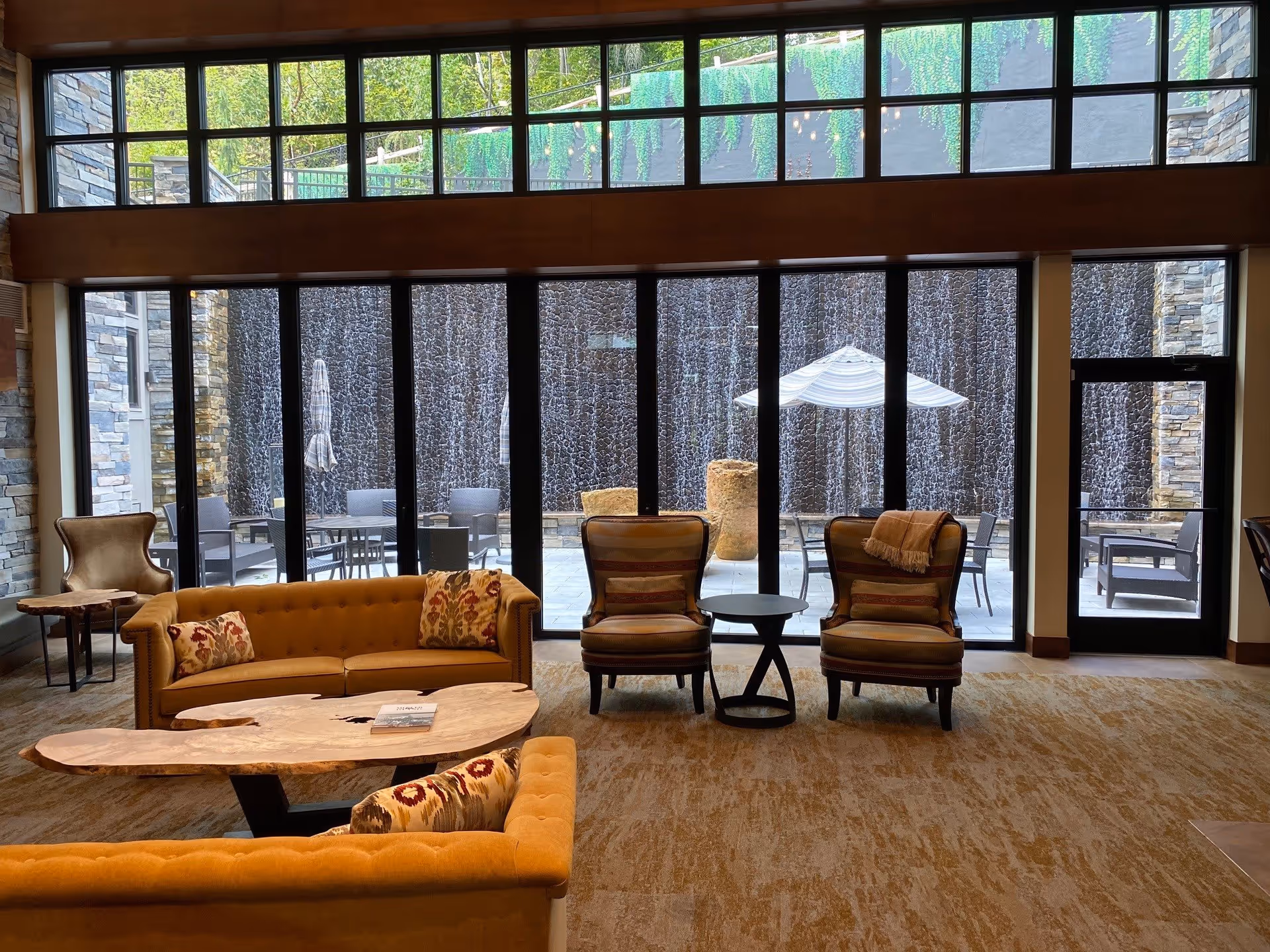 A cozy living room area with mustard yellow sofas and two striped armchairs arranged around a natural wood coffee table. Large floor-to-ceiling windows reveal an outdoor patio with tables, chairs, umbrellas, and a cascading water wall feature. The room has a warm carpet and stone walls on the sides.