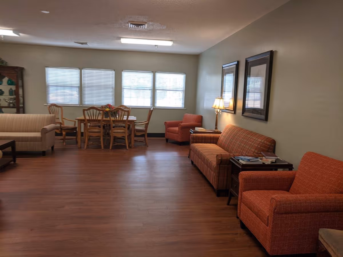 A spacious living room area with wooden flooring, featuring a beige sofa, a wooden dining table with six chairs near windows with blinds, two orange armchairs, an orange plaid sofa, a side table with books and a lamp, and two framed pictures on the wall.
