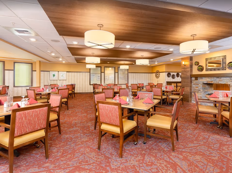 A spacious dining room with multiple wooden tables and chairs arranged neatly. Each table is set with red folded napkins and upside-down glasses. The room features warm lighting with several ceiling lamps, a patterned red carpet, and a stone fireplace on the right side. The walls are decorated with framed artwork and decorative plates.