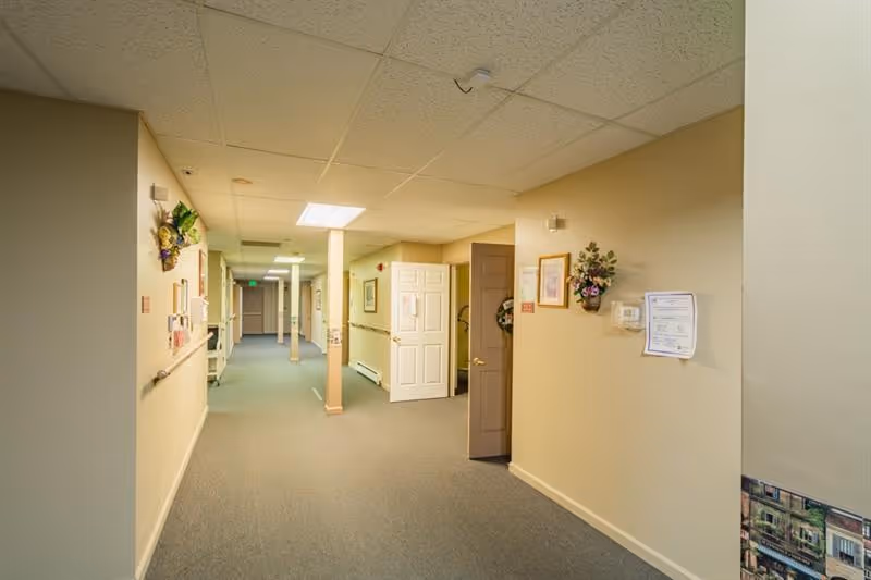 A long carpeted hallway in a senior living facility with beige walls, handrails, doors, and wall decorations.