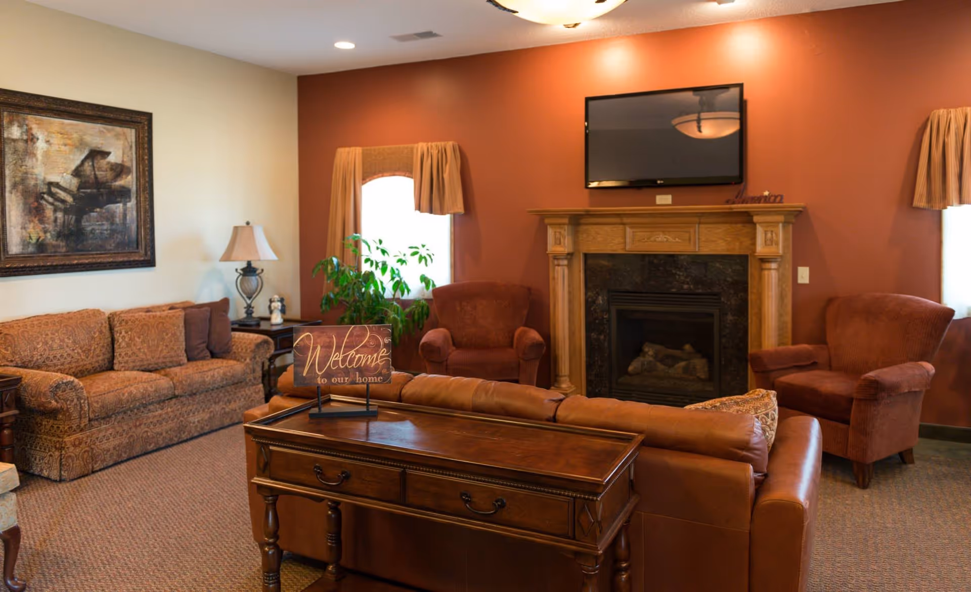 A cozy living room with a brown leather sofa, two upholstered armchairs, and a patterned fabric couch. There is a wooden coffee table with a 'Welcome to our home' sign on it. The room features a fireplace with a wooden mantel and a flat-screen TV mounted above it. Two windows with beige curtains flank the fireplace, and a framed painting of a piano hangs on the wall. A table lamp and a green plant are also visible.